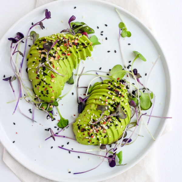 Two halves of an avocado cut into slices on a white plate with micro greens and sesame seeds for garnish.