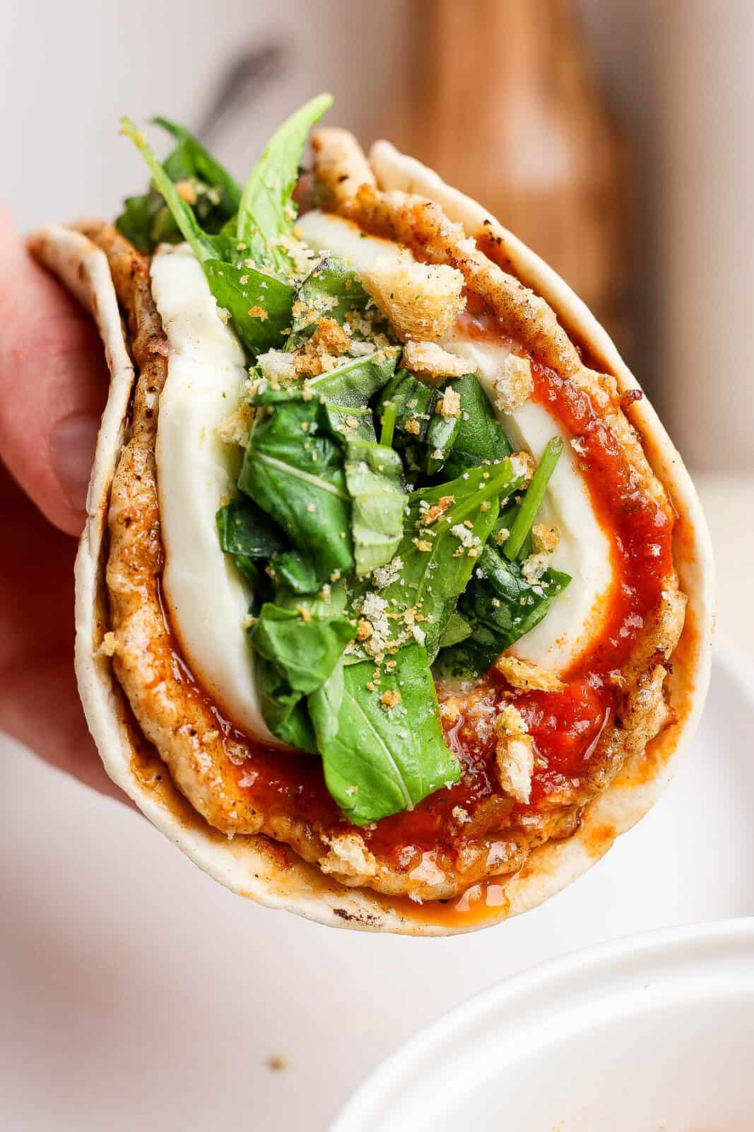 A hand holding a chicken parm smash taco that is topped with an arugula salad and crushed croutons.