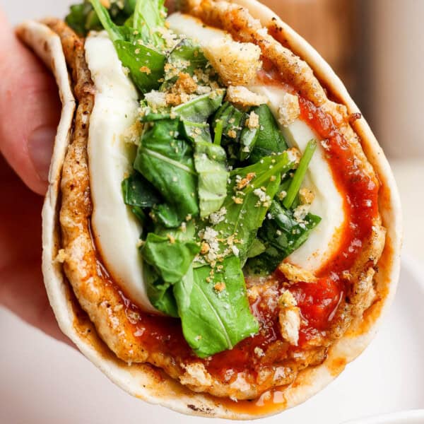 A hand holding a chicken parm smash taco that is topped with an arugula salad mixture and crushed breadcrumbs.
