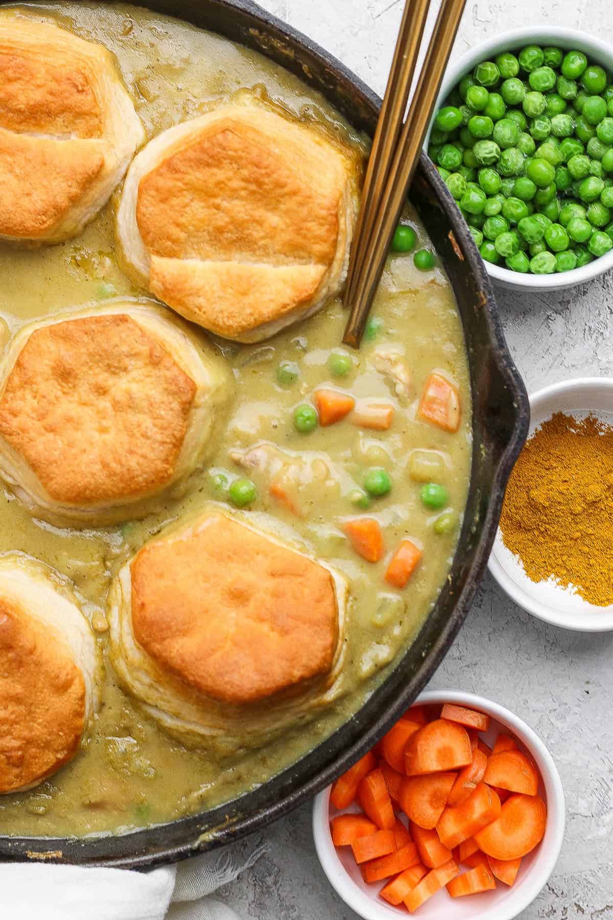 Chicken curry pot pie in a skillet with a spoon next to bowls of peas, curry powder, and carrots.