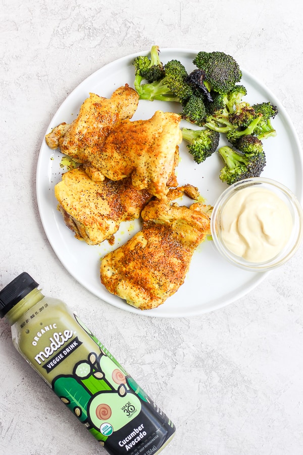 Stove top chicken thighs on a white plate with roasted broccoli, a small bowl of dipping sauce, and a bottle of Medlie veggie drink on the side.