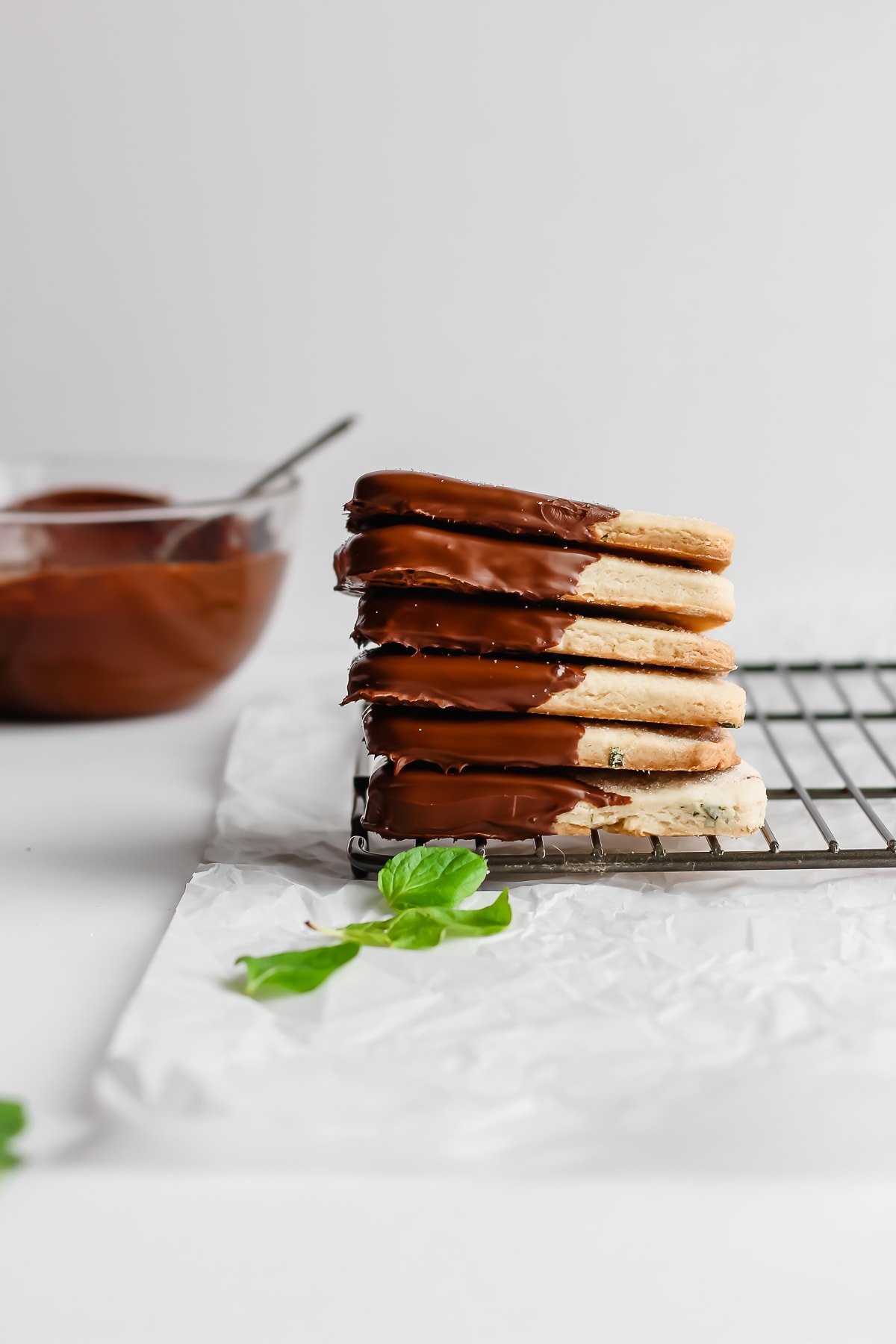 A stack of mint chocolate vegan shortbread cookies on a wire rack next to a bowl of melted chocolate.