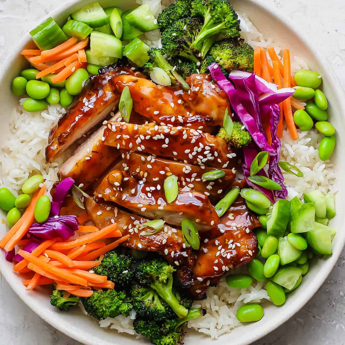 A teriyaki chicken rice bowl with grilled teriyaki chicken sliced over rice with carrots, grilled broccoli, edamame and green onions.