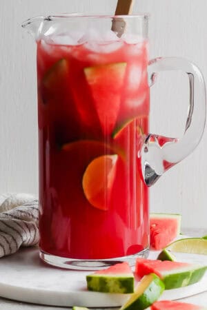 A pitcher of watermelon margaritas with ice, limes and watermelon slices inside and a spoon sticking out.